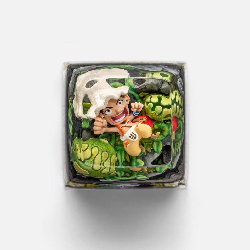 May include: A custom keycap featuring a cartoon character in a dynamic pose, surrounded by green foliage and a white skull-like object. The keycap has a clear resin base, and the character is wearing orange and yellow clothing.