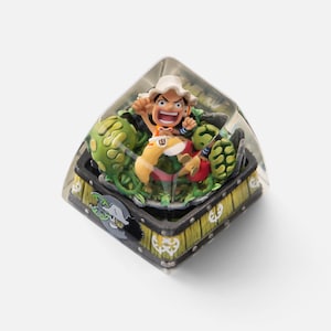 May include: A custom keycap featuring a character from the One Piece anime series. The keycap is encased in a clear resin, with a detailed miniature figure of the character surrounded by green foliage and a wooden chest design.