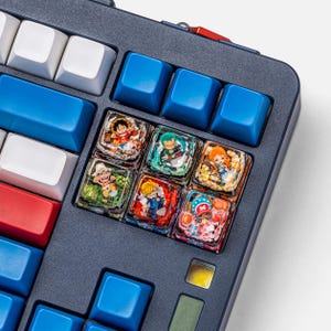 May include: Close-up of a mechanical keyboard with custom keycaps. The keyboard features blue, white, and red keys, with a dark gray frame. Several keycaps are transparent, showcasing colorful anime character designs. The keyboard has an orange accent.