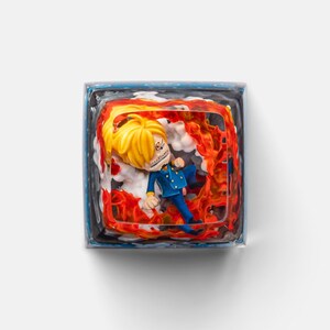 May include: A keycap featuring a character with blond hair and a blue suit, surrounded by flames. The keycap is encased in a clear, cube-shaped resin with a blue and white border, creating a 3D effect. The character appears to be from an anime.