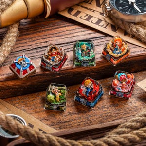 May include: A collection of custom keycaps featuring colorful anime character designs. The keycaps are square and made of clear resin, with detailed figures inside. The designs include a skull and crossbones, and various anime characters. They are displayed on a wooden surface.