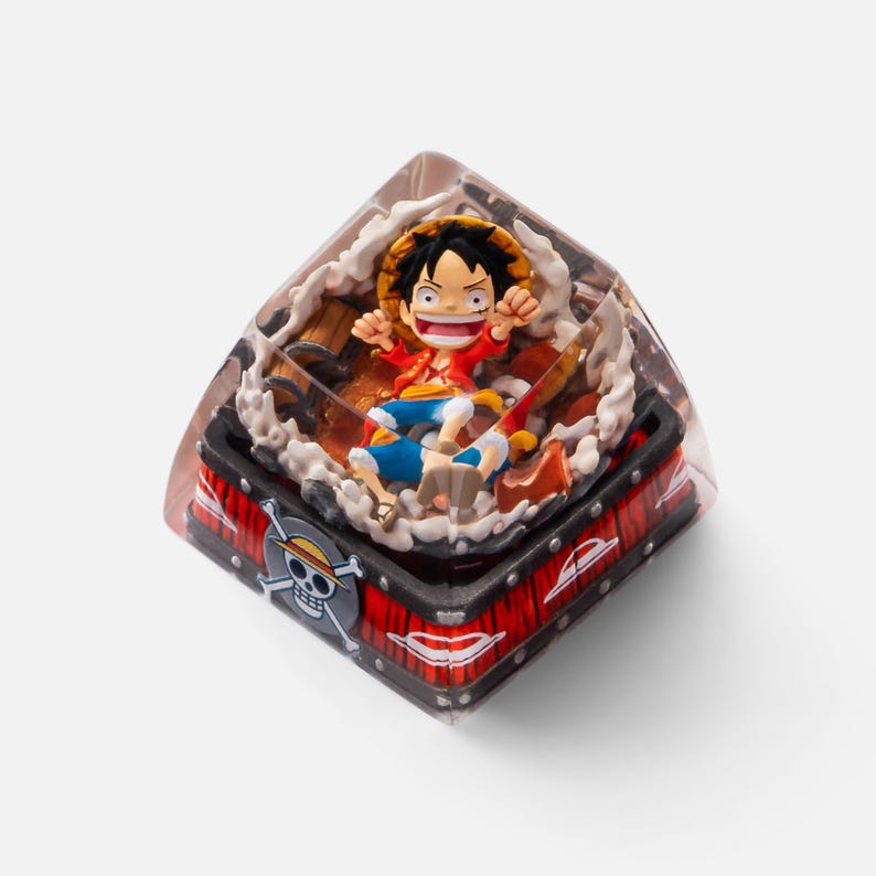 May include: A custom keycap featuring a character from the anime series One Piece. The keycap has a clear resin exterior with a detailed interior scene of the character Luffy. The base is red and black with a skull and crossbones design.