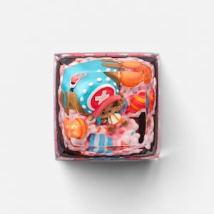 May include: A square, resin keycap featuring a colorful design. The keycap showcases a character with a blue hat and a cross symbol, surrounded by pink and orange elements. The keycap is designed for mechanical keyboards.