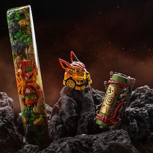 May include: A tall rectangular resin keycap featuring a detailed forest scene with rocks and green foliage, a fox-like character keycap with orange and yellow tones, and a cylindrical scroll keycap with green, gold, and red rope accents.