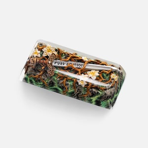 May include: A rectangular resin keycap featuring a miniature sword with runic inscriptions embedded within a scene of brown tangled vines, white flowers, and a dark green textured base. The clear resin casing provides a smooth, glossy finish.