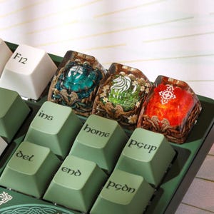 May include: Close-up of a keyboard with custom keycaps. The keycaps feature intricate designs with teal, green, and red accents. The keyboard keys are green with white lettering. The keycaps have a fantasy theme.