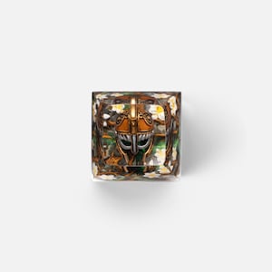 May include: A square resin mechanical keyboard keycap featuring a central bronze-colored helmet with silver accents, surrounded by a detailed background of green foliage and small white flowers encased within the clear, polished material.