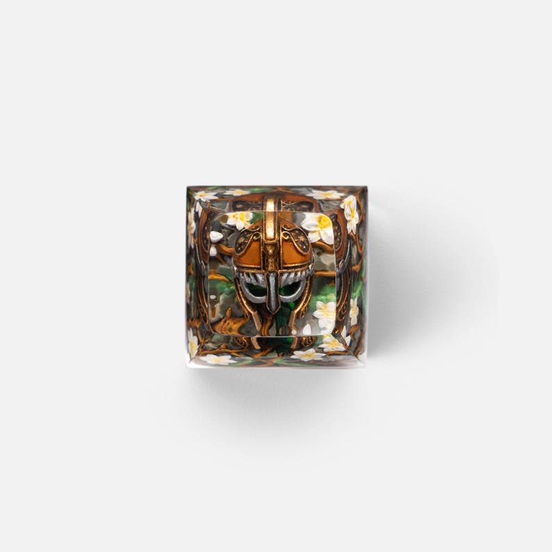 May include: A square resin mechanical keyboard keycap featuring a central bronze-colored helmet with silver accents, surrounded by a detailed background of green foliage and small white flowers encased within the clear, polished material.