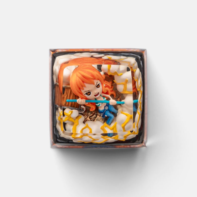 May include: A detailed, square-shaped figurine featuring a character with orange hair and a blue outfit, holding a blue staff. The figure is set within a decorative frame with a cloud-like design and yellow accents. The overall aesthetic is vibrant and detailed.
