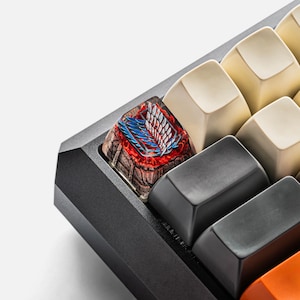 May include: A custom artisan mechanical keyboard keycap featuring a detailed graphic of the Wings of Freedom emblem in blue and white, set against a textured, dark brown stone-like base with vibrant red accents, positioned on a black keyboard.