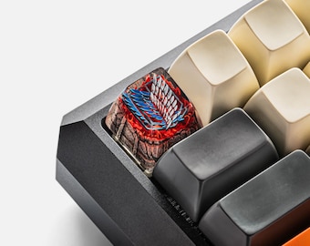 May include: A custom artisan mechanical keyboard keycap featuring a detailed graphic of the Wings of Freedom emblem in blue and white, set against a textured, dark brown stone-like base with vibrant red accents, positioned on a black keyboard.