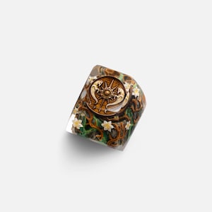 May include: A clear resin ring featuring an embedded circular wooden medallion with a compass design and two horse figures. The band is decorated with brown intertwined branches, green foliage, and small white flowers set within the transparent material.