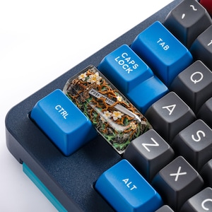 May include: A custom artisan keycap featuring a detailed miniature sword embedded in a clear resin block, surrounded by brown vines and white flowers. The keycap is positioned on a mechanical keyboard between blue CTRL and CAPS LOCK keys.