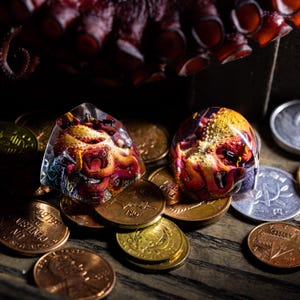 May include: Two clear dice with an octopus design inside. The dice are red, orange, and yellow with a purple and blue background. The dice are sitting on a pile of copper and gold coins.