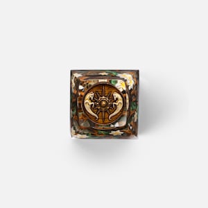 May include: A square resin keycap featuring a central circular emblem with a bronze-colored compass rose and two stylized horse figures. The surrounding clear resin body displays a detailed embedded design of brown branches, green leaves, and small white flowers.