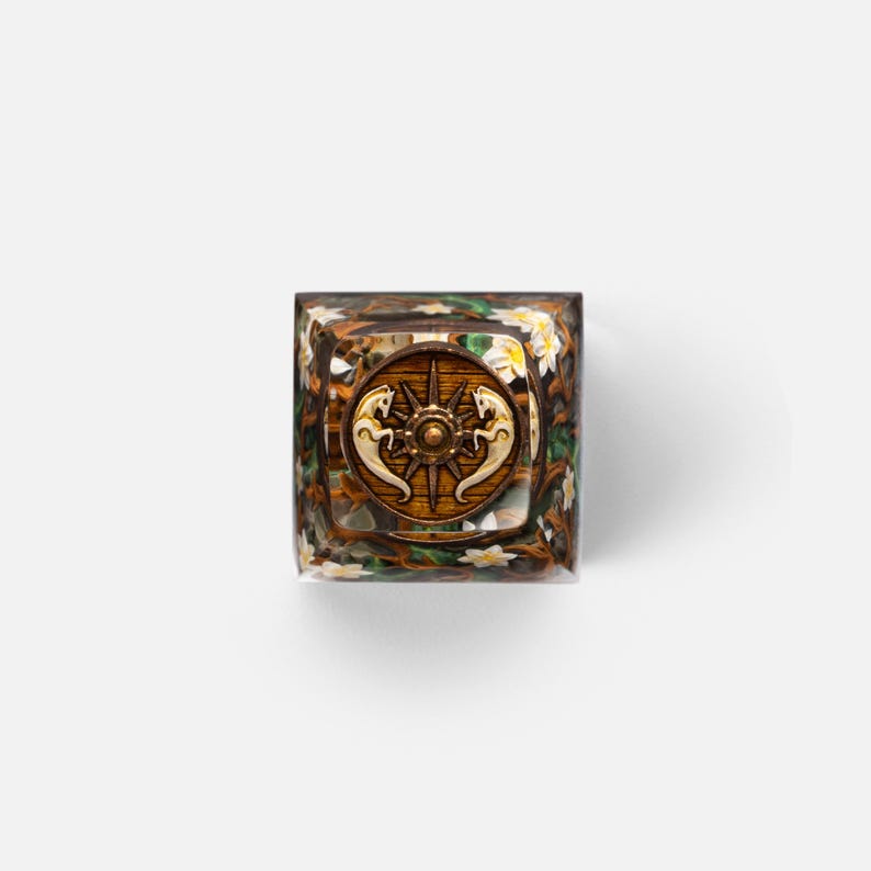 May include: A square resin keycap featuring a central circular emblem with a bronze-colored compass rose and two stylized horse figures. The surrounding clear resin body displays a detailed embedded design of brown branches, green leaves, and small white flowers.