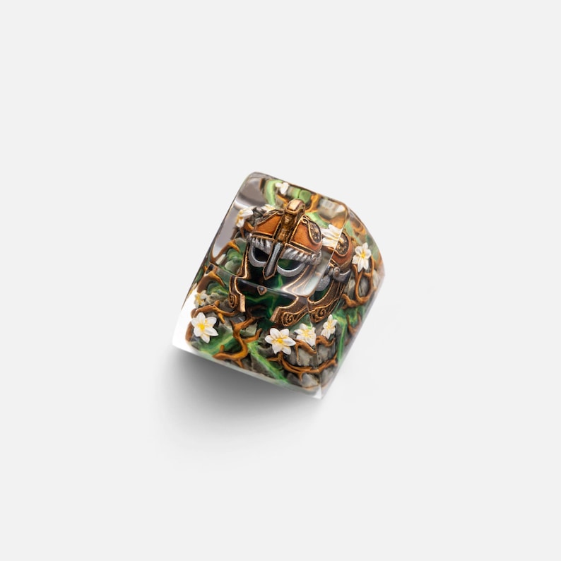 May include: A clear resin mechanical keyboard keycap featuring an intricate, miniature bronze-colored helmet surrounded by green vines and small white flowers. The detailed design is encased within the transparent, cube-shaped artisan keycap.
