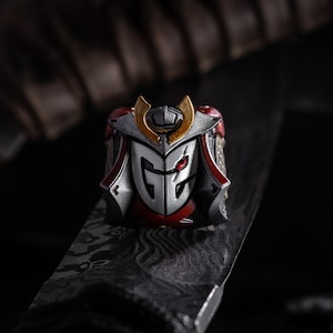 May include: A detailed, silver-colored ring with red and gold accents. The ring features a stylized samurai helmet design with a red eye and the letters "GE" on the front. The ring is set against a dark background.