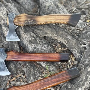 May include: Three hand-forged axes with wooden handles. The top axe has a dark wood handle with a burned design and the number "20" engraved on the head. The middle axe has a reddish-brown handle and the bottom axe has a brown handle.