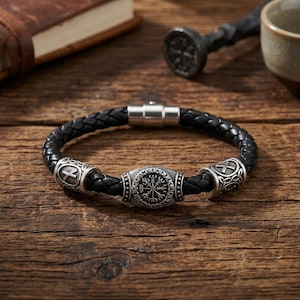 Può includere: Un bracciale in pelle intrecciata nera con una perla centrale in metallo argentato incisa con un simbolo Vegvisir, affiancata da due perle più piccole in metallo argentato con motivi incisi e una chiusura magnetica cilindrica.