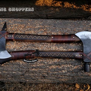 May include: Two Viking choppers, a type of axe, with brown leather handles and a wood shaft. The handles are decorated with a geometric pattern. The axes are laid out on a wooden surface. The text "VIKING CHOPPERS" is visible at the top of the image. The text "Damascus Maker" is visible at the bottom of the image.