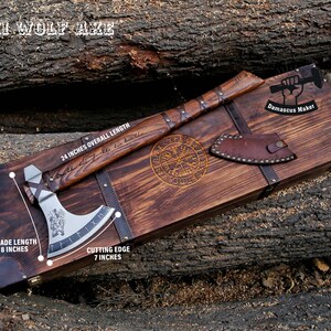 May include: A wooden box with a brown leather handle and a metal axe with a wolf design. The axe is 24 inches long, with an 8-inch blade and a 7-inch cutting edge. The box has the text "Damascus Maker" on it.