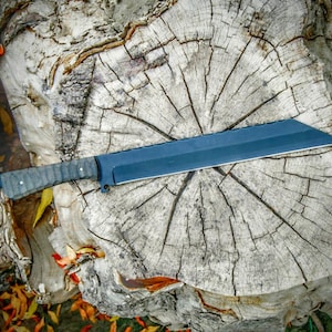 May include: A large, dark blue machete with a textured handle rests on a weathered tree stump. The blade is rectangular with a sharp edge. The handle is a mix of gray and black. The background includes autumn leaves and tree bark.
