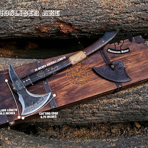 May include: A handcrafted axe with a dark blade and wooden handle, resting on a wooden box. The axe has a 10.5-inch blade and a 6.75-inch cutting edge. The overall length is 26 inches. The box has a decorative design and the text "WINDSLICER AXE".