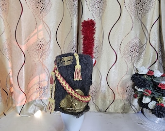 May include: A dark gray military-style hat with a red plume, gold and red tassels, and a gold emblem. The hat has the words "Antique Maker" on it. The hat is displayed on a white mannequin head.