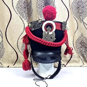 Può includere: Un cappello militare nero con un pompon rosso, corda rossa e nappe. Il cappello ha un bordo in pelle e un distintivo con le parole "Antique Maker". Il cappello è esposto su un supporto bianco.