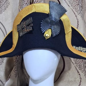 Puede incluir: Un sombrero bicornio negro con ribete dorado y el texto "Antique Maker". El sombrero presenta un emblema decorativo y se exhibe en una cabeza de maniquí blanca. Es una pieza de vestuario histórico.