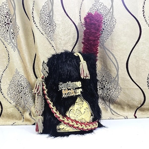 Può includere: Un cappello di pelliccia nera con un pennacchio bordeaux. Il cappello presenta dettagli dorati, nappe e un emblema decorativo con la scritta "Antique Maker". Un cordoncino intrecciato rosso e oro aggiunge dettagli.