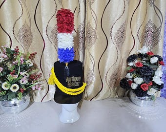 May include: A black hat with a red, white, and blue plume, yellow tassel, and the text "Antique Maker". The hat is displayed on a white stand, with floral arrangements on either side.