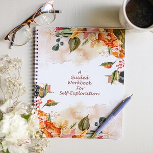 Puede incluir: Un cuaderno blanco de espiral con un diseño floral en la portada. La portada presenta flores de acuarela en tonos de rosa, naranja y amarillo. El texto "A Guided Workbook for Self-Exploration" está impreso en rojo en la portada.