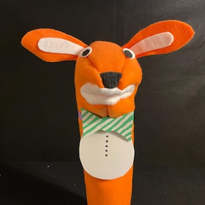 May include: Orange felt hand puppet with white felt ears, a white felt mouth, and a black felt nose. The puppet is wearing a green and white striped bow tie and a white circle with black dots.
