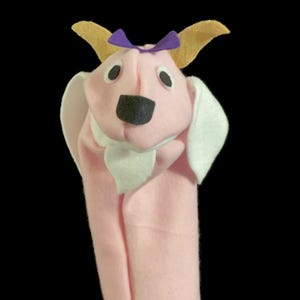 Può includere: Marionetta a mano in feltro rosa con un colletto in feltro bianco, un naso in feltro nero e orecchie in feltro giallo. La marionetta ha un fiocco in feltro viola sulla testa.