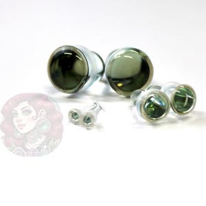 Op de afbeelding: Een verzameling glazen oordopjes en oorbellen. De pluggen zijn groot, met een donkergroen midden en heldere randen. De oorbellen zijn kleiner, met een vergelijkbare groene tint. Op een witte ondergrond.