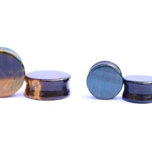 May include: Two pairs of stone ear plugs. One pair features a mix of brown and gold tones, while the other pair has blue and black hues. The plugs have a rounded, flat face and a flared, concave back.