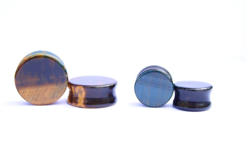 May include: Two pairs of stone ear plugs. One pair features a mix of brown and gold tones, while the other pair has blue and black hues. The plugs have a rounded, flat face and a flared, concave back.
