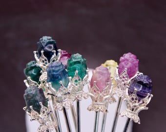 May include: A collection of gemstone hair sticks, each featuring a carved rose design in various colors including green, blue, purple, and pink. The rose heads are mounted on silver-colored metal sticks.