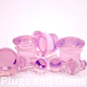 May include: A collection of iridescent pink glass plugs in various sizes. The plugs have a rounded, domed top and a flared base. The text "EC Plugs and Gems" is visible in the lower left corner.