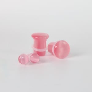May include: Pink cat's eye style ear plugs in various sizes. The plugs have a rounded, smooth surface and a light pink color. The image shows several plugs, some with a flared design and others with a more traditional shape.