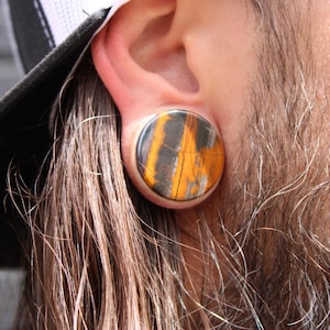 May include: A large, round ear plug with a striking pattern of orange, black, and gray stripes. The plug is inserted into an ear, showcasing the unique design and texture of the jewelry. The plug is a bold accessory.
