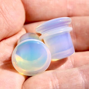 May include: Two opalescent ear plugs with a pearlescent sheen. The plugs have a rounded front and a cylindrical body with a flared edge. The colors shift from white to blue and yellow.