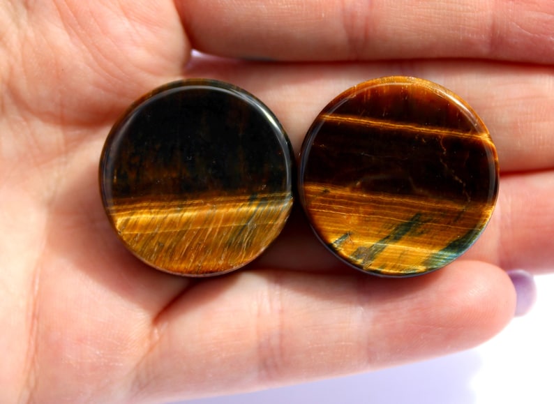 May include: Two round tiger's eye stone plugs. The plugs are held in a hand and have a polished, smooth surface. The colors are a mix of dark brown, gold, and hints of blue-green, with a striped pattern. These are likely used for ear piercings.