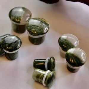 A striking pair of Green Jasper Single Flare Plugs is displayed against an earthy-toned backdrop. Each plug features deep forest green hues with natural patterns of dark veining and subtle swirls, unique to every piece. The surface is smoothly polish