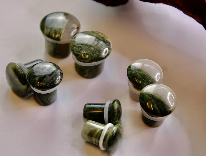 A striking pair of Green Jasper Single Flare Plugs is displayed against an earthy-toned backdrop. Each plug features deep forest green hues with natural patterns of dark veining and subtle swirls, unique to every piece. The surface is smoothly polish