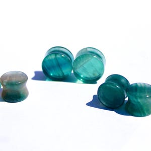 May include: Several teal and green stone ear plugs are arranged on a white surface. The plugs have a flared design and a polished, smooth texture. The stone has a marbled appearance with varying shades of green and teal.