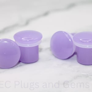 May include: Four lavender-colored ear plugs with a rounded top and cylindrical base. The plugs have a white rim between the top and base. The text "EC Plugs and Gems" is at the bottom of the image.