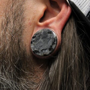 May include: Close-up of a gray and black stone ear plug. The plug is round and set in a flesh-colored ear. The person has a gray beard and long gray hair. A black and white hat is visible.